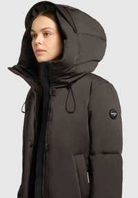Giacca piumino marrone scuro con un grande cappuccio, zip frontale, coulisse regolabile e patch con logo sulla manica sinistra. Tessuto liscio e isolato.