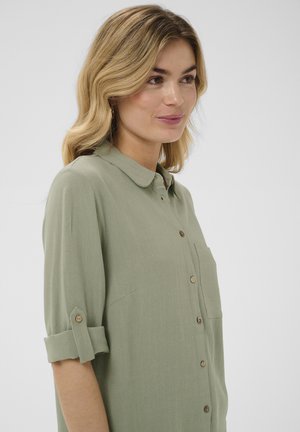 Femme aux cheveux blonds mi-longs portant une chemise vert clair boutonnée avec les manches retroussées, face légèrement tournée vers la droite sur un fond uni.