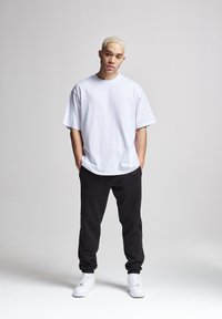 Weißes übergroßes T-Shirt, schwarze Jogginghosen und weiße Sneakers. Schlichtes Design, Baumwollmaterial, stehend auf einer glatten Oberfläche mit neutralem Hintergrund.