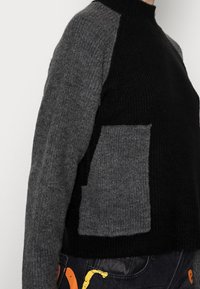 Stickad tröja med hög halsringning, i svart kropp och grå ärmar med en ficka i lappar. Materialet verkar mjukt och texturerat.