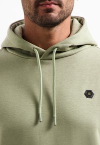 Lichtgroene hoodie gemaakt van zachte stof, met een hexagonaal zwart logoaccent en trekkoorden. Gladde textuur met een ontspannen pasvorm.