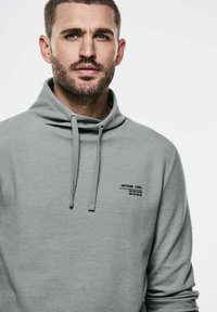 Šedá mikina s vysokým límcem, stahovací šňůrkou a malým černým textem na levé části hrudi. Text obsahuje souřadnice a název značky. Měkká textura.