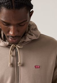 Sweatshirt cu glugă de culoare bej, din material texturat, cu fermoar frontal și șnur. Prezintă un mic logo roșu Levi's pe piept.