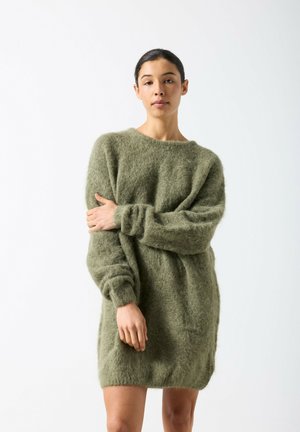 Noor naine, kes seisab käed rüpes, tal on seljas pikk, karvane oliivroheline kampsunist kleit, taust on ühtlaselt valge.