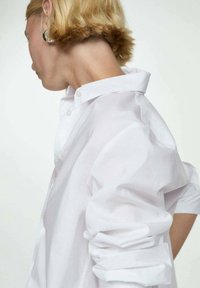 Personne aux cheveux blonds courts porte une chemise blanche boutonnée, tournée vers la gauche avec la tête légèrement inclinée vers le bas sur un fond clair uni.