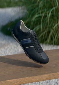 Sötétkék tornacipő sima bőr felsőrésszel, szürke részletekkel, fűzős záródással és mintás gumitalppal, egy fából készült felület felett emelkedve.