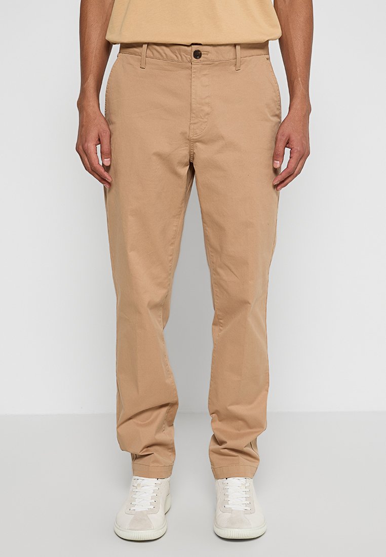 Beige Baumwollhose mit geradem Bein, ausgestattet mit einem Frontknopfverschluss und schrägen Seitentaschen, kombiniert mit weißen Sneakern.