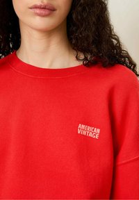 Röd sweatshirt i mjukt tyg, med rund halsringning och texten "AMERICAN VINTAGE" broderad i vitt på bröstet.