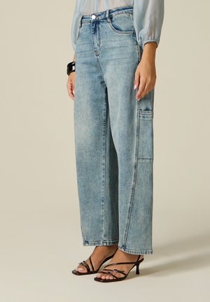 Vrouw draagt lichtblauwe wijdlopende jeans met zijzak en zwarte sandalen met hak en bandjes tegen een neutrale achtergrond.