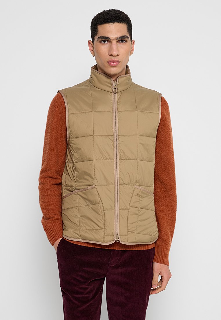 Gilet imbottito beige con colletto alto, motivo trapuntato e due tasche laterali. Chiusura con zip e rifiniture a contrasto. Indossato sopra un maglione ruggine.