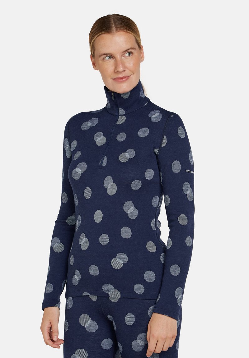 Frau in navybluse Langarmshirt und Hose mit hellgrauen Punkten, steht mit entspannten Händen und schaut nach rechts.