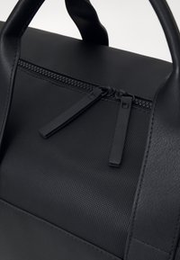 Schwarze strukturierte Tasche mit einem oberen Reißverschluss und flachen Zippergriffen sowie kontrastierenden glatten Lederakzenten an den Griffen und der Seite.
