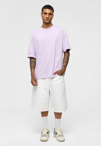 Lichtpaarse oversized t-shirt met verlaagde schouders, gecombineerd met witte shorts tot de knie, witte sokken en beige sneakers met zwarte accenten.