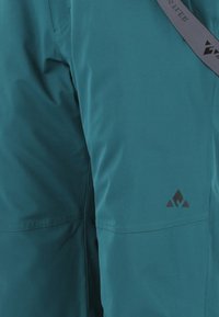 Teal Outdoorhosen mit einem kleinen schwarzen geometrischen Logo am Oberschenkel und einem grauen Riemen mit schwarzem Text, der seitlich herunterhängt.