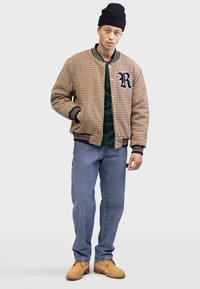 Veste bomber à carreaux marron avec des poignets et un col côtelés, accents navy, chemise verte en dessous, jeans bleus et bottes beiges.
