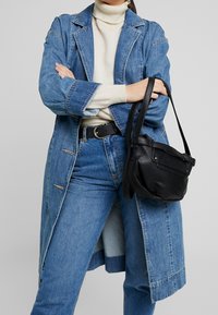 Cappotto lungo in denim, azzurro chiaro, con colletto strutturato e tasche laterali. Borsa a mano in pelle nera con finitura testurizzata e tracolla regolabile.