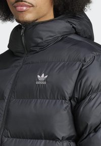 Fekete puffermellény magas gallérral, cipzáras záródással és puha, fényes anyagból készült. Ezüst Adidas logóval rendelkezik a mellkason.