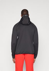 Melna jakas ar kapuci ar gludu faktūru, uz kuras ir logo pie kakla un raglāna piedurknēm, saskaņota ar spilgti sarkanām biksēm.