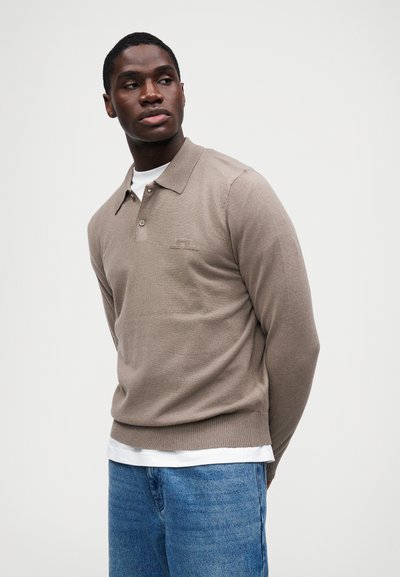 Καστανή πλεκτή πουκαμίσα πόλο με γιακά, ραμμένα μανίκια και στρίφωμα. Περιλαμβάνει ένα διακριτικό λογότυπο στο στήθος, το οποίο είναι τοποθετημένο πάνω από ένα λευκό πουκάμισο και μπλε τζιν.