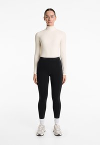 Hosszú ujjú, testhezálló krém színű felső fekete, magas derekú leggingsszel; a modellen világos edzőcipő és fehér bokazokni van.