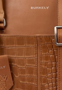 Sac en cuir marron avec texture crocodile, surface lisse et quincaillerie en métal. Présente le nom de la marque embossé en argent, accompagné d'une étiquette.