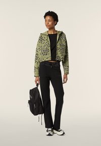 Haut à capuche cropped vert olive avec un imprimé léopard noir et blanc, associé à un pantalon noir et des baskets noir et blanc ; sac à dos noir à la main.