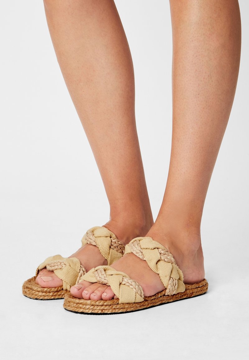 Sandales en tissu beige tressé avec une semelle en jute texturée, présentant un design plat et un bout ouvert, idéales pour une tenue décontractée.