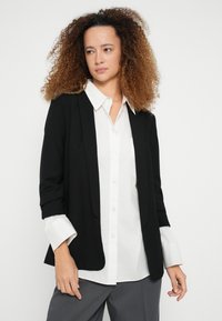 Μαύρο σακάκι με εφαρμοστή γραμμή, μαλακό ύφασμα και γυρισμένα μανίκια πάνω από λευκό πουκάμισο με κουμπιά και γκρι παντελόνι. Απλός και μοντέρνος σχεδιασμός.