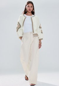 Veste crème brodée avec manches ballon, associée à un pantalon beige à jambes larges et des sandales noires. Chemise blanche simple en dessous.