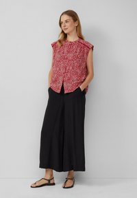 Rode, met patronen bedekte mouwloze top met een ronde halslijn, gecombineerd met zwarte wijde broek en platte sandalen, gesitueerd tegen een neutrale achtergrond.