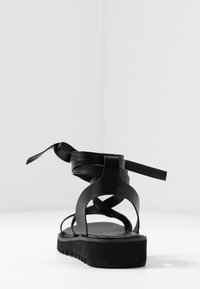 Černé kožené sandály s plochou podrážkou, s křížovými pásky a zadním vázacím detailem. Hladký povrch a minimalistický design.