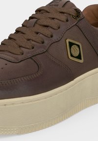 Gros plan d'une basket en cuir marron avec une semelle épaisse couleur crème, des lacets texturés et un emblème de marque or et noir sur le côté.
