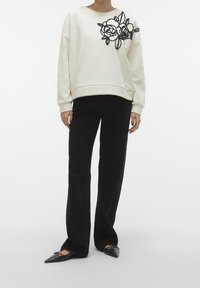 Person wearing a white sweatshirt with black floral embroidery on the shoulder, black wide-leg pants, and black pointed flats.