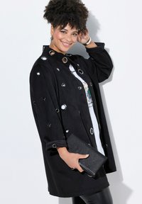 Chaqueta negra con grandes ojales plateados, mangas enrolladas y corte recto. Combinada con un clutch negro texturizado y una camiseta gráfica blanca debajo.