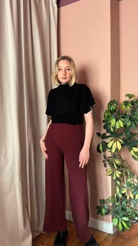 Šviesiaplaukė moteris juodu viršutiniu drabužiu ir bordo plataus kirpimo kelnėmis stovi patalpoje šalia smėlio spalvos užuolaidos ir margalapio žaliuojančio kambarinio augalo.