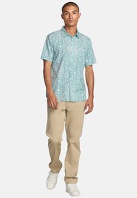 Short-sleeve button-up shirt in teal with white abstract patterns, paired with beige pants and white sneakers. Features a small logo.