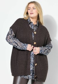 Cardigan marrone lavorato a maglia con maniche corte, caratterizzato da scollo a V, grandi bottoni dorati e un motivo testurizzato. Indossato sopra una camicia blu a fantasia.