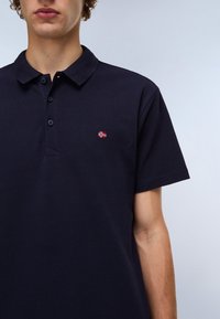 Marineblå pique-skjorte i pustende stoff, med en treknapplukking og et lite broderi av det norske flagget på brystet.