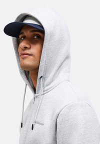 Light gray hooded sweatshirt with adjustable drawstrings and a front zipper. Features a navy cap with a flat brim.