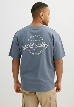 Hombre con el pelo corto y pendiente, vestido con una camiseta azul "Wild Valley Campbase" y pantalones cortos cargo beige, de espaldas frente a un fondo liso.