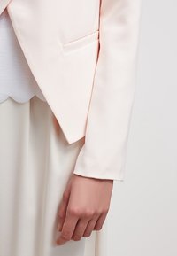 Jasnoróżowa marynarka o dopasowanym kroju, gładka w dotyku, z jedną kieszenią z przodu, zestawiona z białą bluzką z falbaniastym wykończeniem, częściowo widoczną.