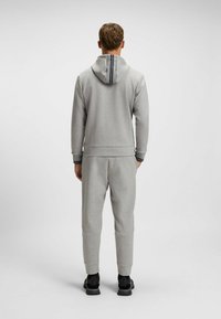 Felpa grigia con cappuccio regolabile, con un'accortezza a strisce. Abbinata a pantaloni da tuta tapered coordinati e scarpe da ginnastica nere.