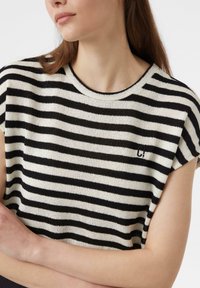 Gestreifter Strickoberteil in Creme und Schwarz, mit kurzen Ärmeln und rundem Halsausschnitt. Verfügt über ein kleines Logo auf der Brust.