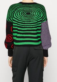 Pull à rayures vertes et noires avec un motif en spirale, accents rouges et une manche texturée violette. Poignets et ourlet côtelés.