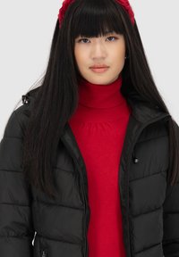 Giacca piumino nera con un design trapuntato, dotata di cappuccio e tasche con zip, indossata sopra un maglione a collo alto rosso.