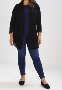 Cardigan leggero nero con maniche a tre quarti, con dettagli in pizzo all'orlo, indossato sopra un top blu navy e abbinato a jeans skinny blu e ballerine nere.