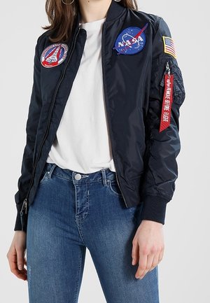 Person trägt eine schwarze NASA-Bomberjacke mit Aufnähern, ein weißes Hemd und blaue Jeans und steht vor einem schlichten Hintergrund.