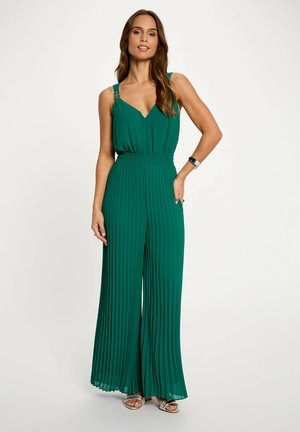 Donna in piedi con la mano in tasca, indossa una tuta verde senza maniche plissettata e sandali argento con punta aperta su uno sfondo semplice.