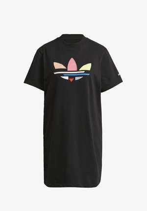 Kobieta ubrana w czarną sukienkę z krótkim rękawem z kolorowym logo Adidas oraz białe buty sportowe Adidas i paskowane skarpetki do kostki, stojąca.