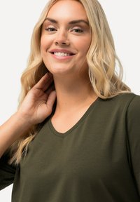 Olijfgroene V-hals top van zachte stof, met driekwartmouwen en een gladde textuur. Model heeft lang blond haar.